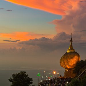 Golden Rock Pagoda in Myanmar glowing at dusk on a cliffside, with pilgrims below and a colorful sunset sky filled with dramatic clouds.