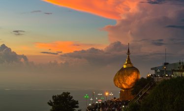 Golden Rock Pagoda in Myanmar glowing at dusk on a cliffside, with pilgrims below and a colorful sunset sky filled with dramatic clouds.