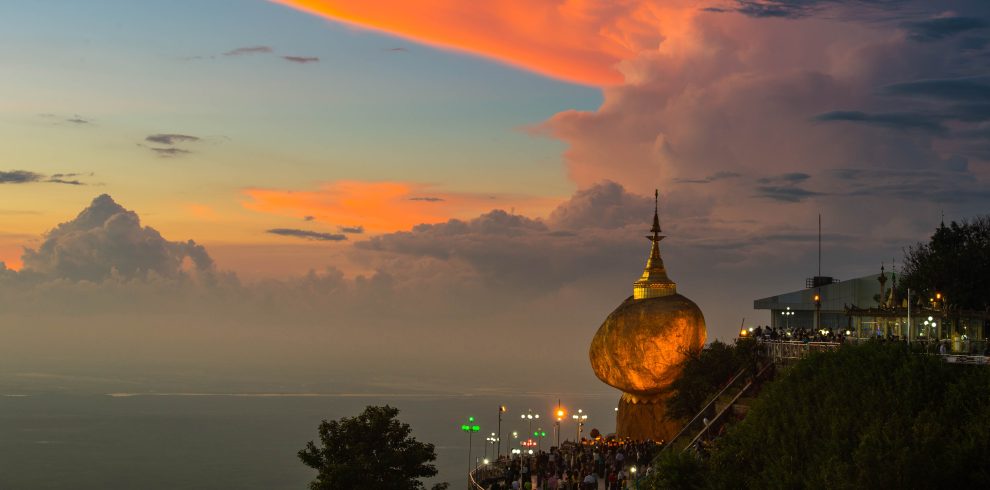 Golden Rock Pagoda in Myanmar glowing at dusk on a cliffside, with pilgrims below and a colorful sunset sky filled with dramatic clouds.