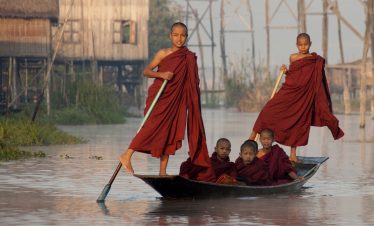 Young Buddhist monks standing on a wooden boat collecting alms on Inle Lake, Myanmar, at sunrise.