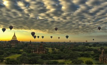 Hot air balloons floating over the temples and pagodas of Bagan at sunrise beneath a dramatic cloud-filled sky.