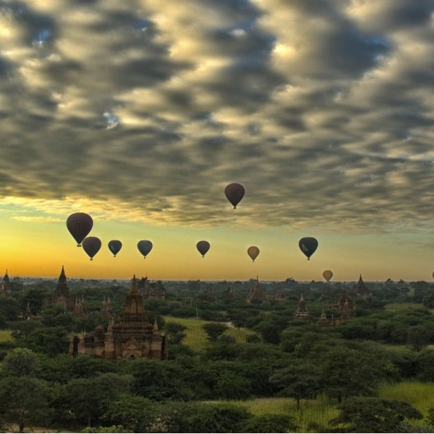 Hot air balloons floating over the temples and pagodas of Bagan at sunrise beneath a dramatic cloud-filled sky.