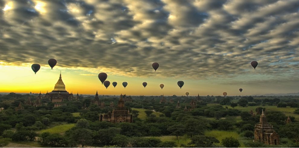 Hot air balloons floating over the temples and pagodas of Bagan at sunrise beneath a dramatic cloud-filled sky.