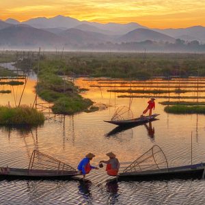 Traditional leg-rowing fishermen on Inle Lake at sunrise, boats gliding through floating gardens with misty mountains in the background.