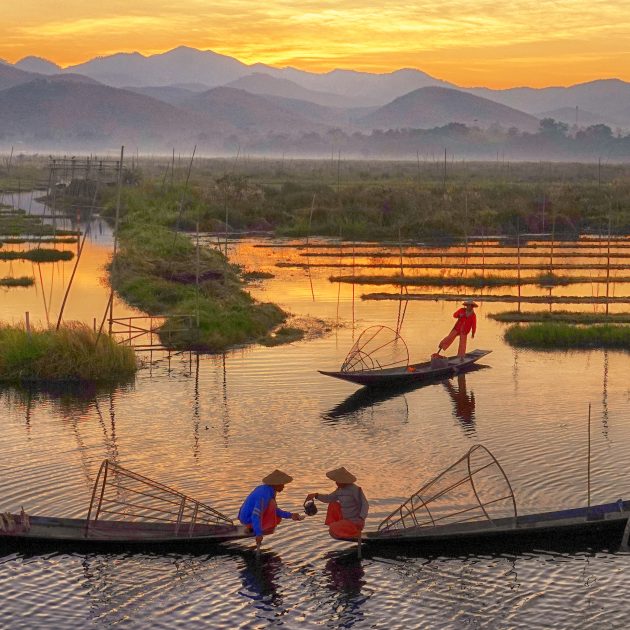 Traditional leg-rowing fishermen on Inle Lake at sunrise, boats gliding through floating gardens with misty mountains in the background.
