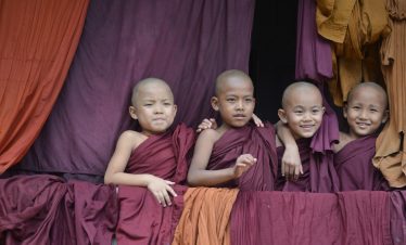 Group of young novice monks from Myanmar smiling and leaning on a ledge, framed by maroon and saffron robes.
