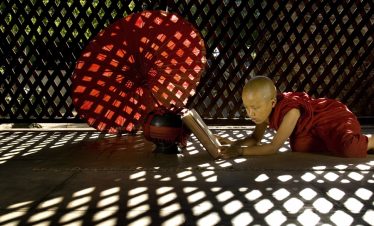 Young Buddhist novice in a red robe reading a book on the floor beside a red parasol, lit by patterned light through a lattice wall.