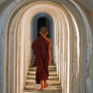 Young novice monk in a maroon robe walking barefoot through a tunnel of repeating white arches.