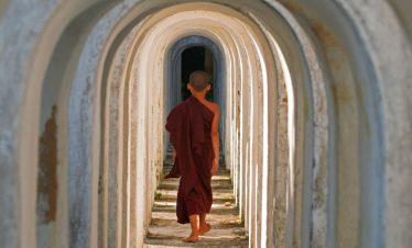 Young novice monk in a maroon robe walking barefoot through a tunnel of repeating white arches.