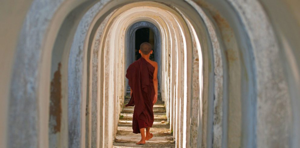 Young novice monk in a maroon robe walking barefoot through a tunnel of repeating white arches.