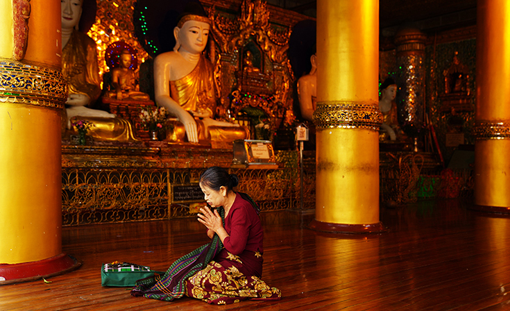 Woman kneeling in prayer inside Shwedagon Pagoda surrounded by golden Buddha statues in Yangon, Myanmar.