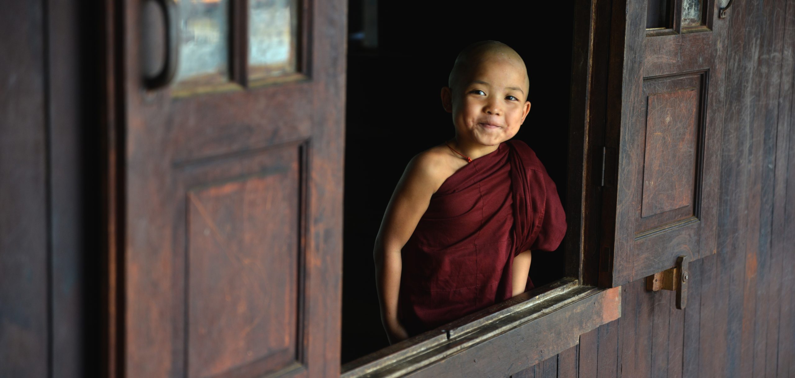 Young novice monk smiling through a wooden monastery window in Myanmar