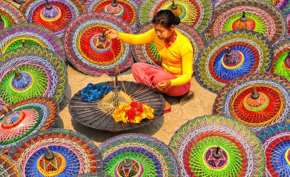 Woman in bright clothes weaving threads on a traditional parasol, surrounded by colorful circular umbrellas.