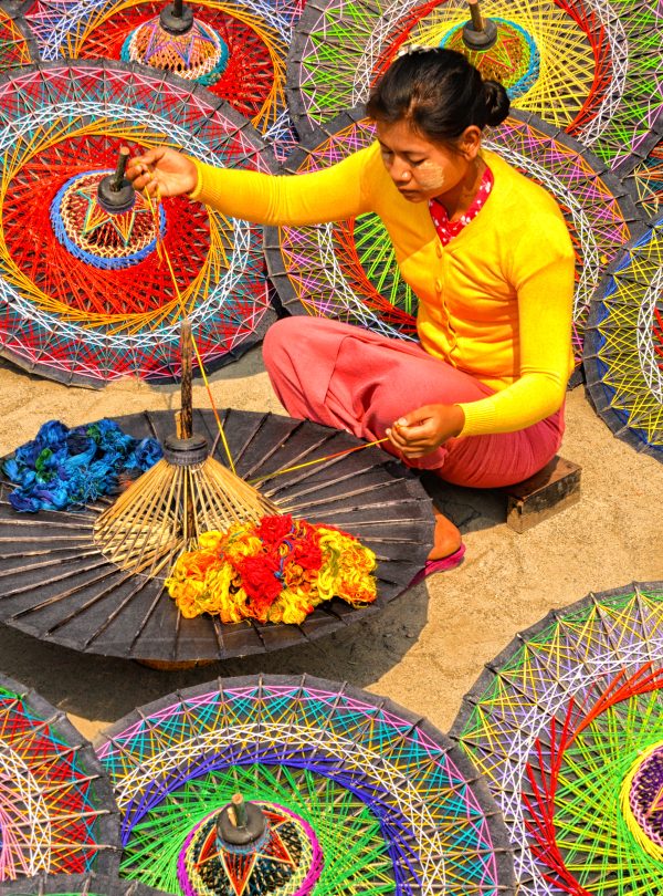 Woman in bright clothes weaving threads on a traditional parasol, surrounded by colorful circular umbrellas.