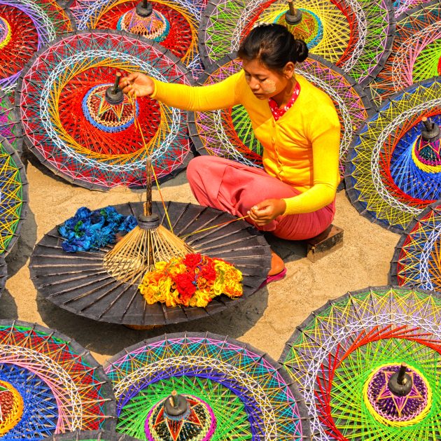 Woman in bright clothes weaving threads on a traditional parasol, surrounded by colorful circular umbrellas.