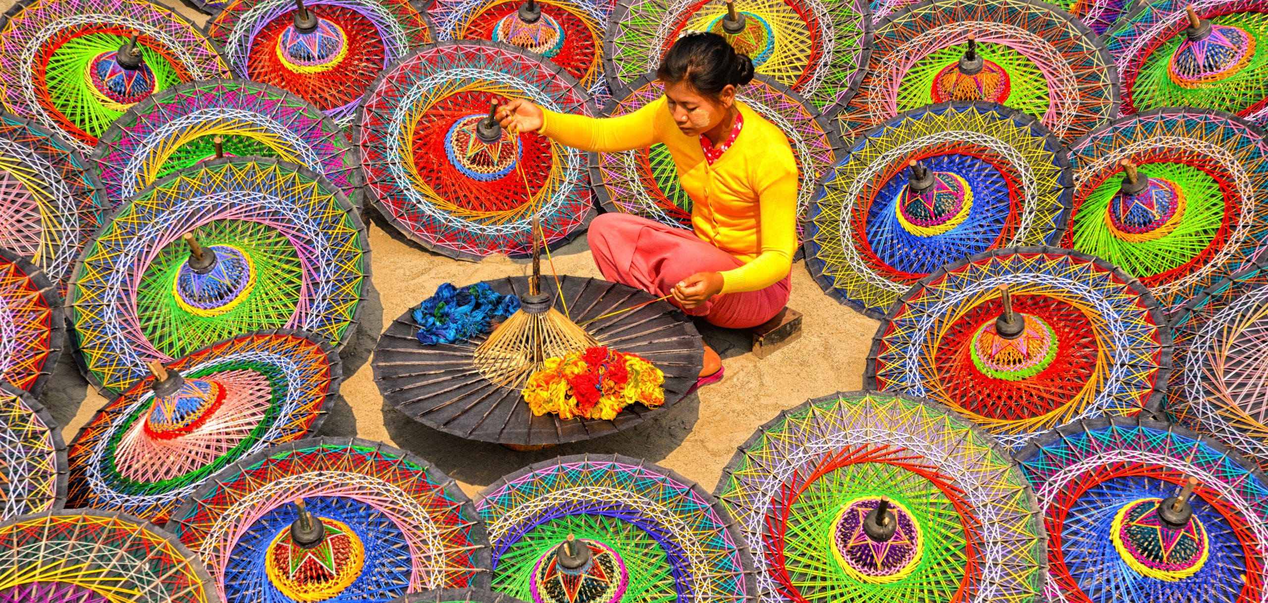 Woman in bright clothes weaving threads on a traditional parasol, surrounded by colorful circular umbrellas.
