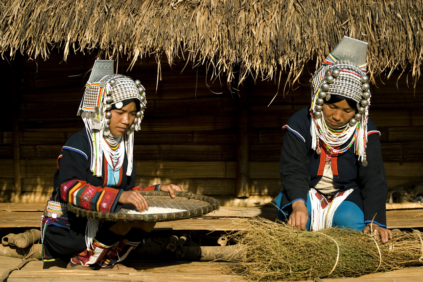 Hill-tribe women in ornate traditional headdresses winnowing rice and sorting herbs outside a thatched house in Myanmar.