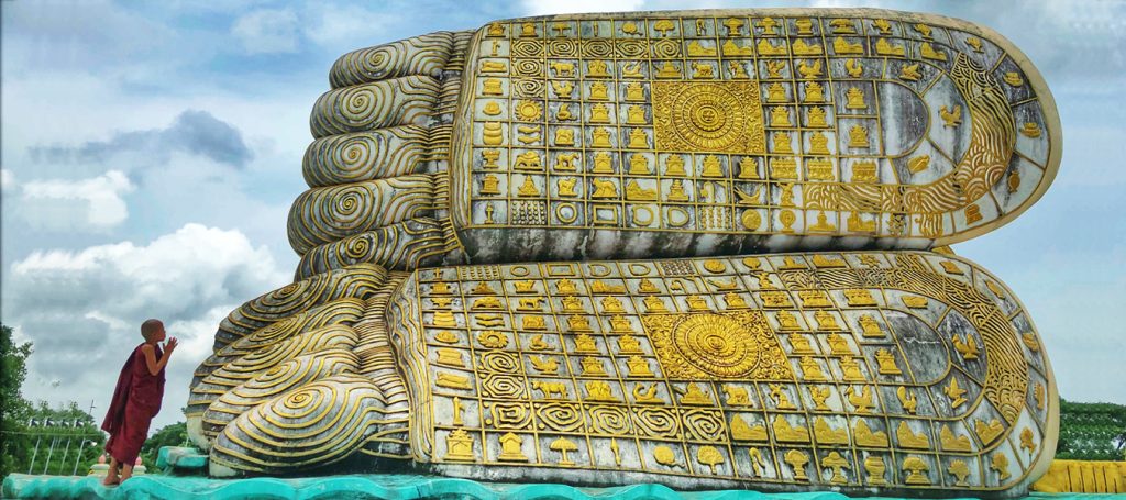 Close-up of the gilded carved soles of a reclining Buddha statue with a young novice monk praying beside them under a bright sky.