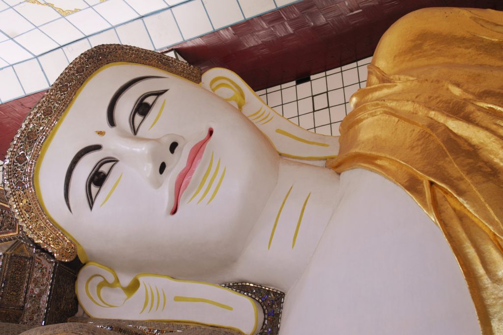 Close-up of the serene face of a gilded reclining Buddha statue with an ornate headpiece and golden robe inside a Myanmar temple.