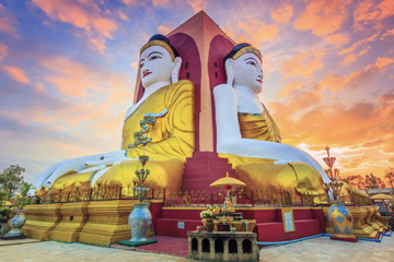 The four seated Buddhas of Kyaikpun in Bago, Myanmar, shown back-to-back on a decorated pedestal with a colorful sunset sky.