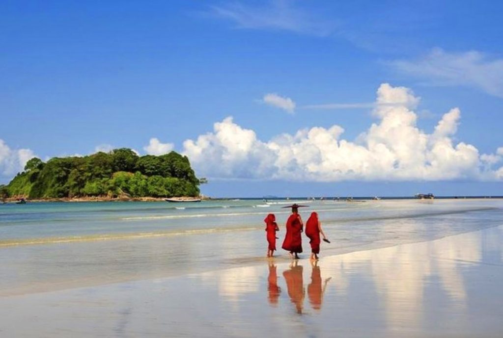Three Buddhist monks in red robes walking along the wet sand of Ngwesaung Beach, Myanmar, with a small green island and blue sky reflected on the shoreline.