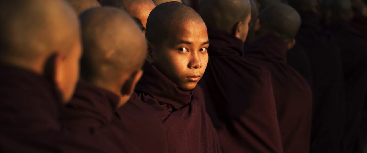 Young novice monk in maroon robes, lit by warm temple light, looking toward the camera while standing in a line of fellow monks in Myanmar.