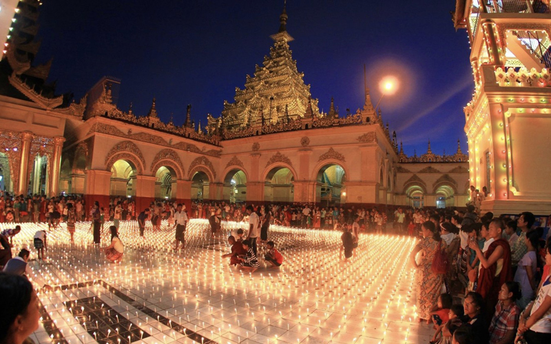 Thousands of candles fill a temple courtyard at night as crowds gather for the Festival of Lights (Thadingyut) with the illuminated Shwedagon Pagoda in the background.