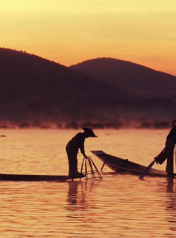 Silhouettes of Inle Lake leg-rowing fishermen lifting cone nets at sunrise with misty hills in the background — Inle Lake, Myanmar.