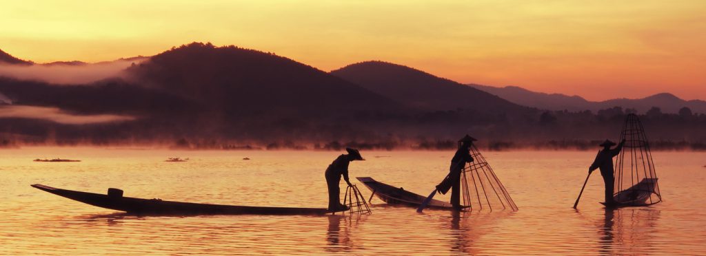 Three Intha fishermen using the traditional leg-rowing technique on long wooden boats at dawn on Inle Lake, with a conical fishing net and soft reflections on the water.