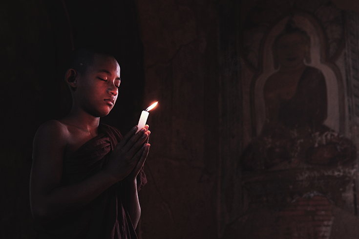 Young novice monk holding a lit candle while praying inside an ancient temple in Bagan, Myanmar.