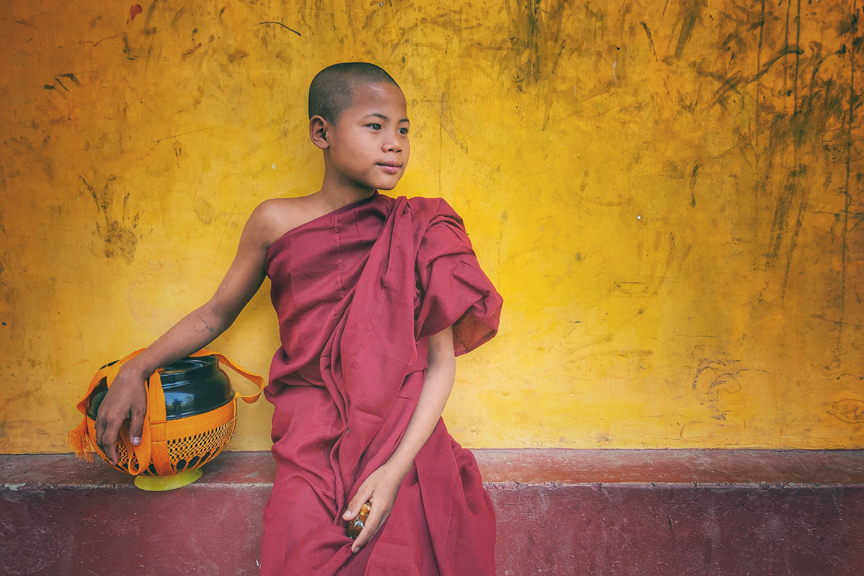 Young novice Buddhist monk in maroon robes sitting against a weathered yellow temple wall with his alms bowl beside him.