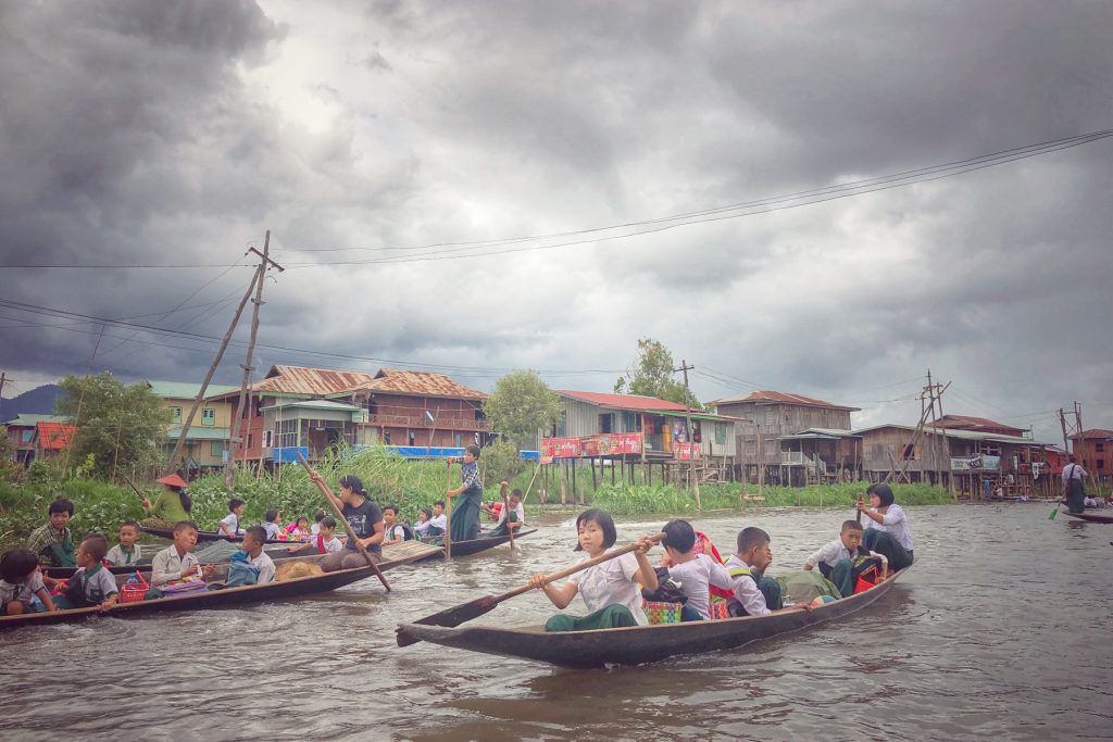 Schoolchildren and villagers riding long wooden boats past stilt houses in a floating village on Inle Lake, Myanmar, under a dramatic cloudy sky.