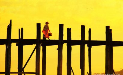 Nun walking across U Bein Bridge at sunrise in Amarapura, Mandalay, Myanmar, silhouetted against a bright golden sky.