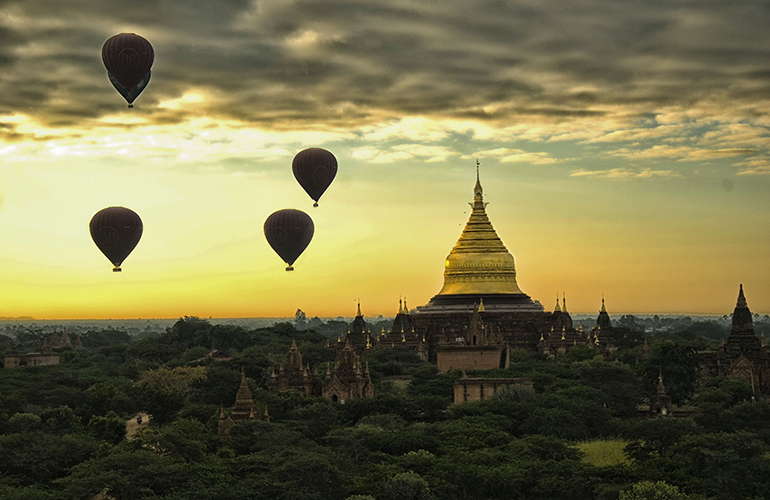 Hot air balloons flying above ancient temples at sunrise in Bagan, Myanmar with a golden pagoda in the background.