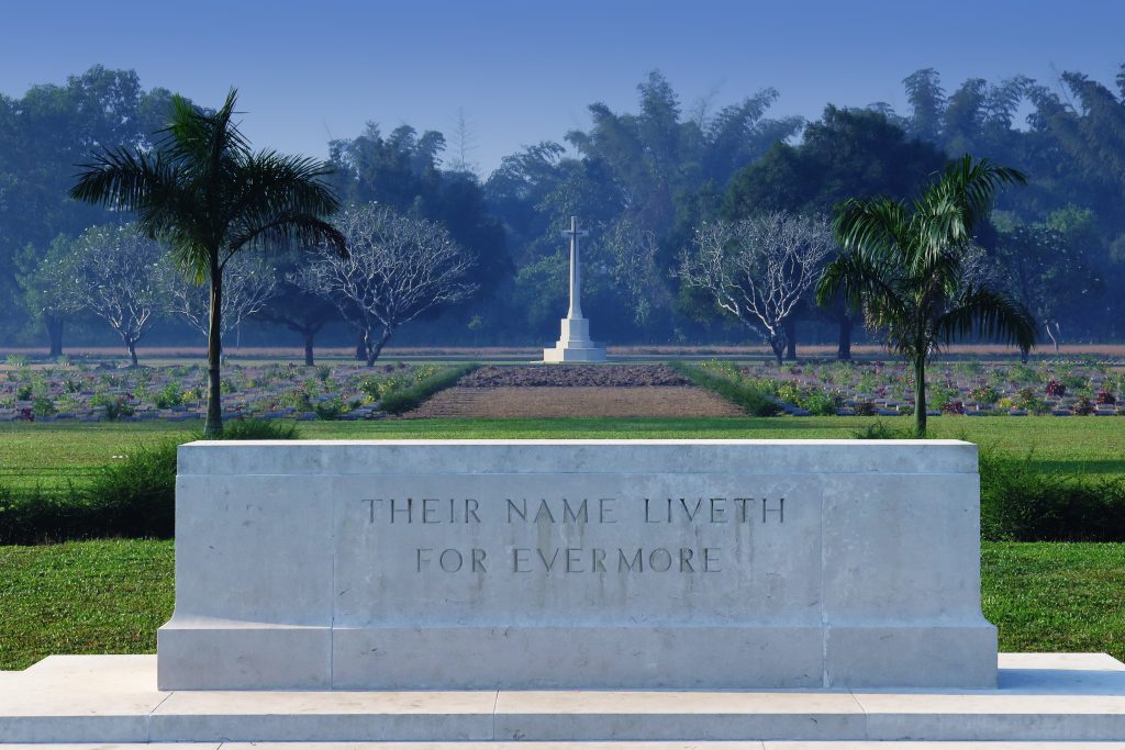 Stone memorial engraved with “THEIR NAME LIVETH FOR EVERMORE” in the foreground, with a cross and neat rows of war graves stretching across a green cemetery under a misty tree line.