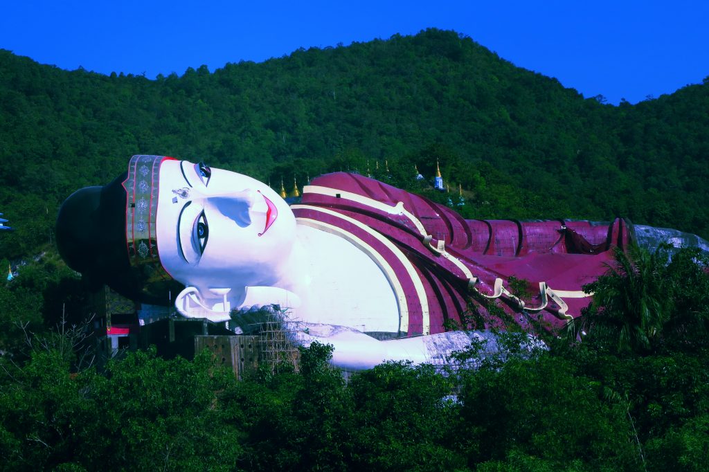 Large reclining Buddha statue with a white face and maroon-striped robe resting on a green hillside in Myanmar.