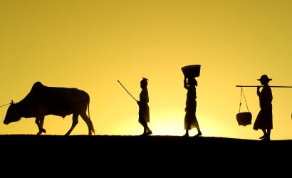 Silhouettes of villagers and an ox walking along a ridge at sunrise, carrying baskets and poles in rural Myanmar.
