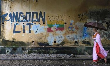 Buddhist nun in pink and red robes walks with an open umbrella along a rocky railway track beside a weathered wall covered in graffiti reading “Rangoon City.”