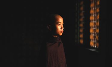 Young Buddhist novice in a dark robe stands in soft window light, looking to the side against a dark background.