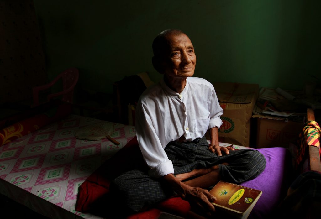 Elderly Myanmar man sitting indoors in soft window light holding a book