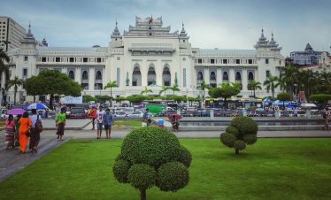 People with umbrellas walk through a green park toward Yangon City Hall, a large white colonial-style building with tiered Burmese roofs, palm trees, and busy traffic in front.
