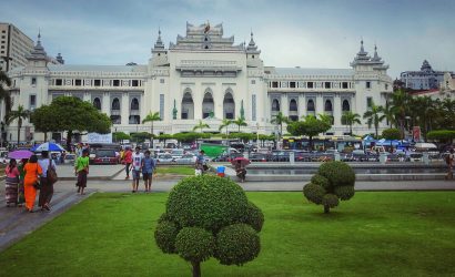 People with umbrellas walk through a green park toward Yangon City Hall, a large white colonial-style building with tiered Burmese roofs, palm trees, and busy traffic in front.