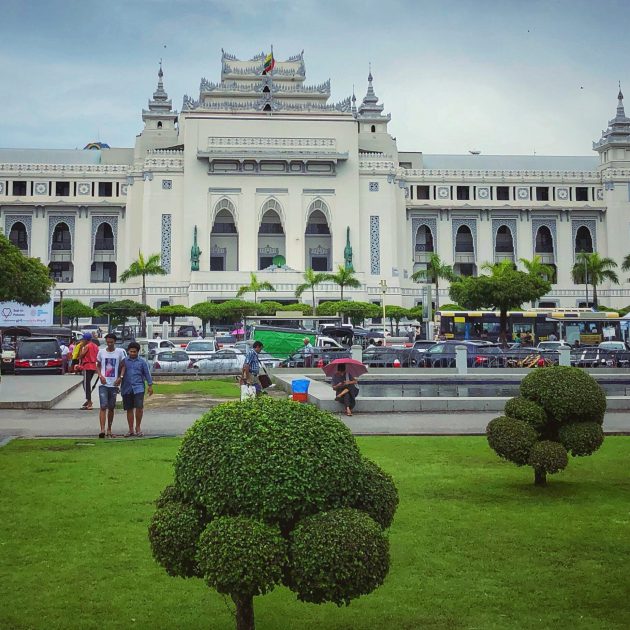 People with umbrellas walk through a green park toward Yangon City Hall, a large white colonial-style building with tiered Burmese roofs, palm trees, and busy traffic in front.