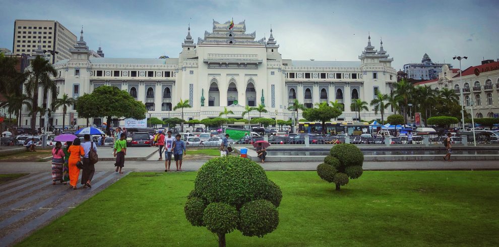 People with umbrellas walk through a green park toward Yangon City Hall, a large white colonial-style building with tiered Burmese roofs, palm trees, and busy traffic in front.