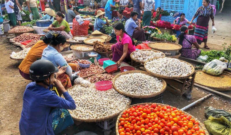 Vendors selling vegetables, garlic, tomatoes, and fresh produce at a busy outdoor market in Yangon, Myanmar.